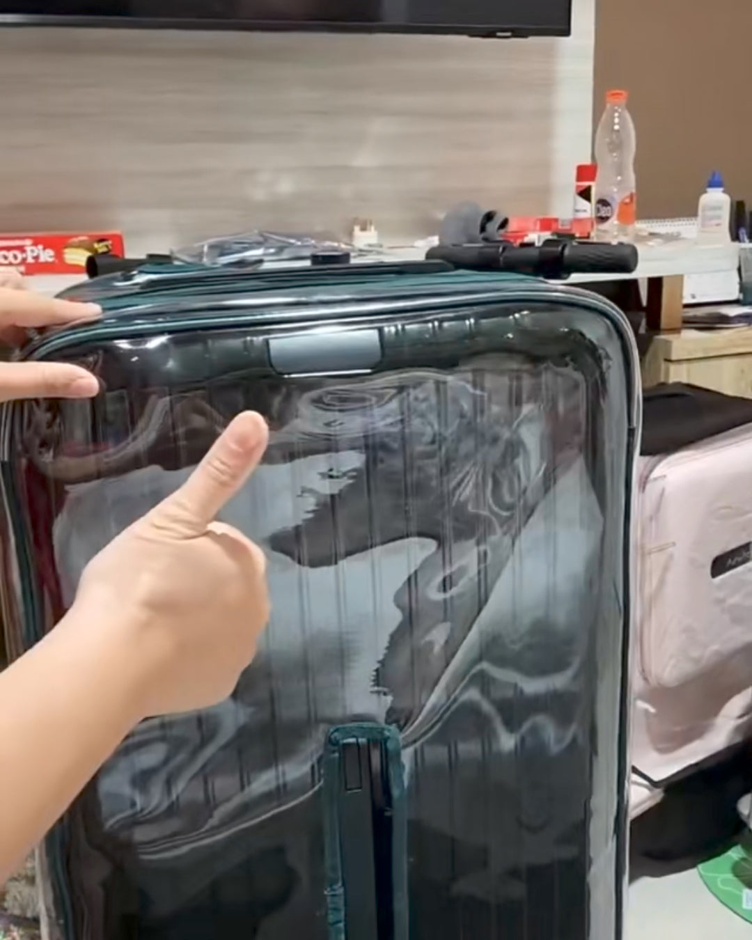 Rimowa Essential Trunk Plus in a clear Fixmeuniva cover with green trim, with a hand giving a thumbs-up beside it.